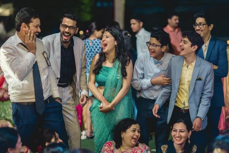 A group of corporate employees in formal and semi-formal attire, including a woman in a green saree and men in blazers, laughing together during a comedy-themed evening event.
