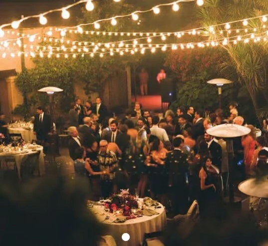 A crowded outdoor corporate event at night with a forest theme, featuring warm string lights draped across the trees, round dining tables with floral centerpieces, and guests in formal attire.