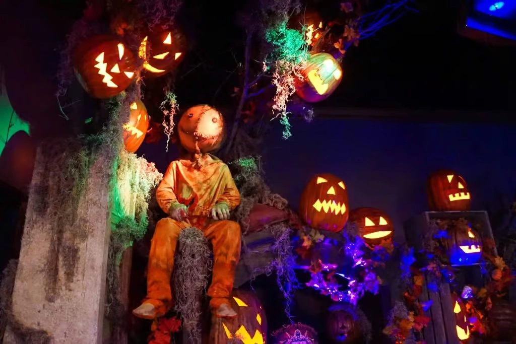 A spooky corporate event setting featuring a scarecrow-like figure with a pumpkin head, surrounded by glowing jack-o'-lanterns and eerie green and purple lighting.