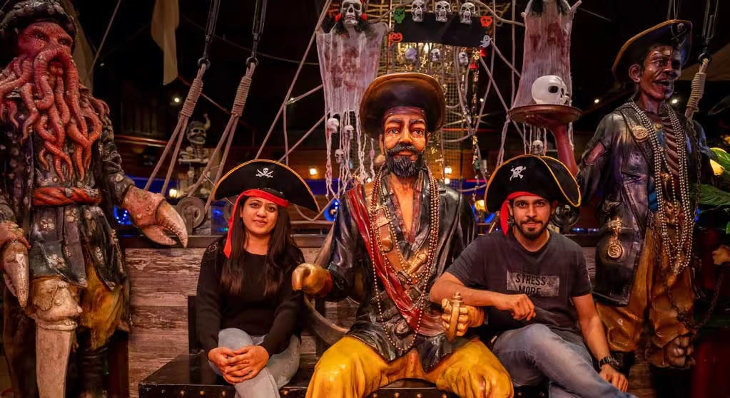 Two corporate employees wearing pirate hats posing with life-sized, highly detailed pirate statues including a captain and a Davy Jones-like figure in an eerie, thematic setting.