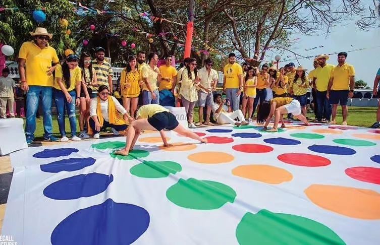 A group of corporate employees in matching yellow t-shirts playing a giant version of Twister on a large outdoor lawn during a sports-themed event.