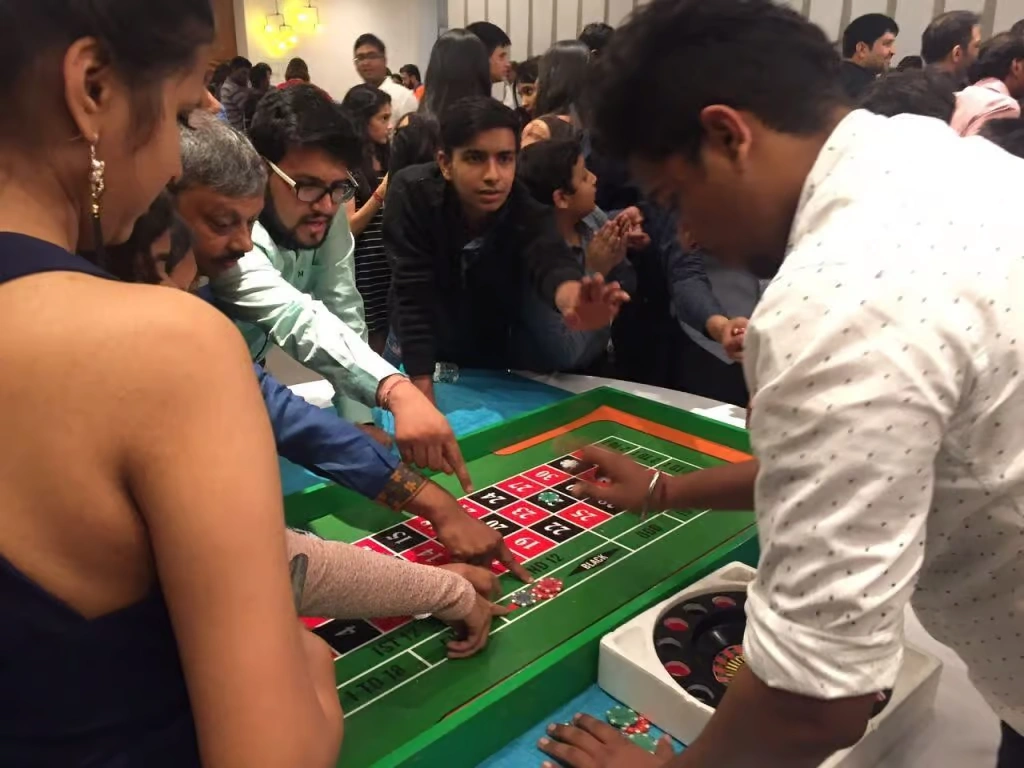 A group of corporate employees gathered around a green roulette table, actively placing bets with red and black chips during a themed casino night event.