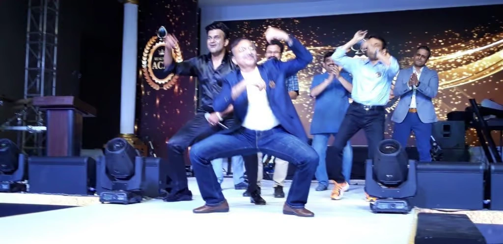 A group of six corporate employees dressed in vibrant 70s and 80s retro attire, including a red afro wig, neon pink LED glasses, and vintage blue-tinted sunglasses, posing together in a brightly lit event hall.