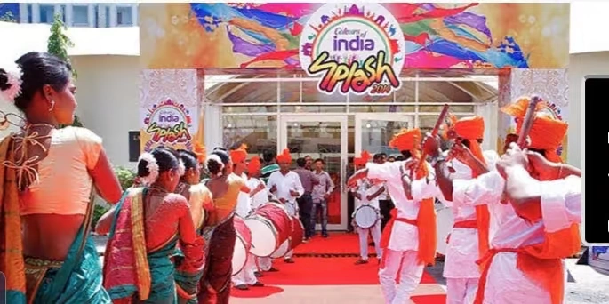 A grand entrance to a corporate event titled "Colours of India Splash," featuring a red carpet, traditional Dhol players in white and orange attire, and women in green and gold sarees welcoming guests.