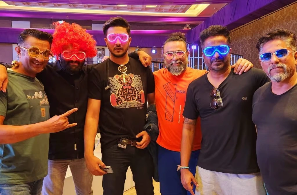 A group of corporate employees dressed in vibrant 70s and 80s retro attire, including polka dots, headbands, and vintage prints, posing together in a brightly lit event hall.