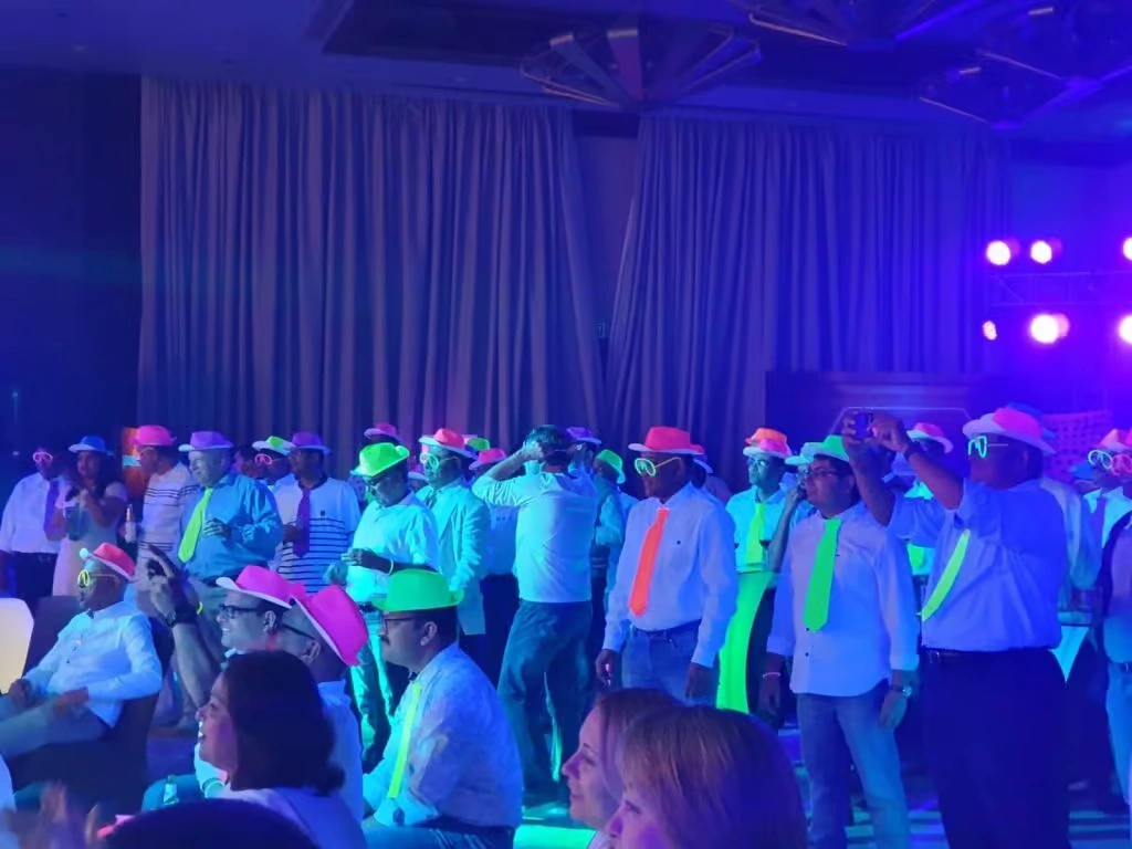 A group of corporate employees wearing glowing neon hats, LED glasses, and fluorescent ties dancing under UV blacklights in a purple-lit event hall.