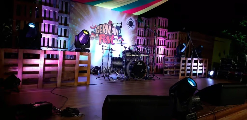 A professional event stage at night featuring a "German Fest" backdrop with a drum set, surrounded by walls of stacked wooden pallets, vibrant purple and yellow stage lighting, and colorful draped fabric overhead.