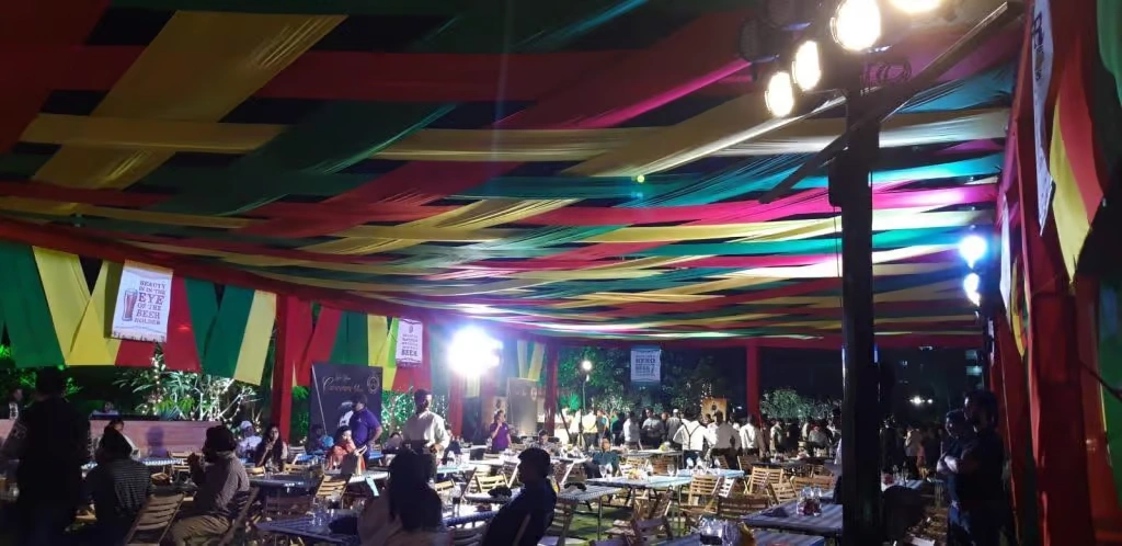 A large outdoor corporate event space under a canopy of crisscrossing yellow, red, and green fabric strips, featuring long communal tables, warm ambient lighting, and groups of people networking in a festival-style atmosphere.