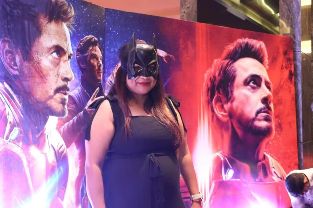 A corporate employee wearing a black dress and a Batman mask standing in front of a large Marvel-themed backdrop featuring Iron Man and Doctor Strange.