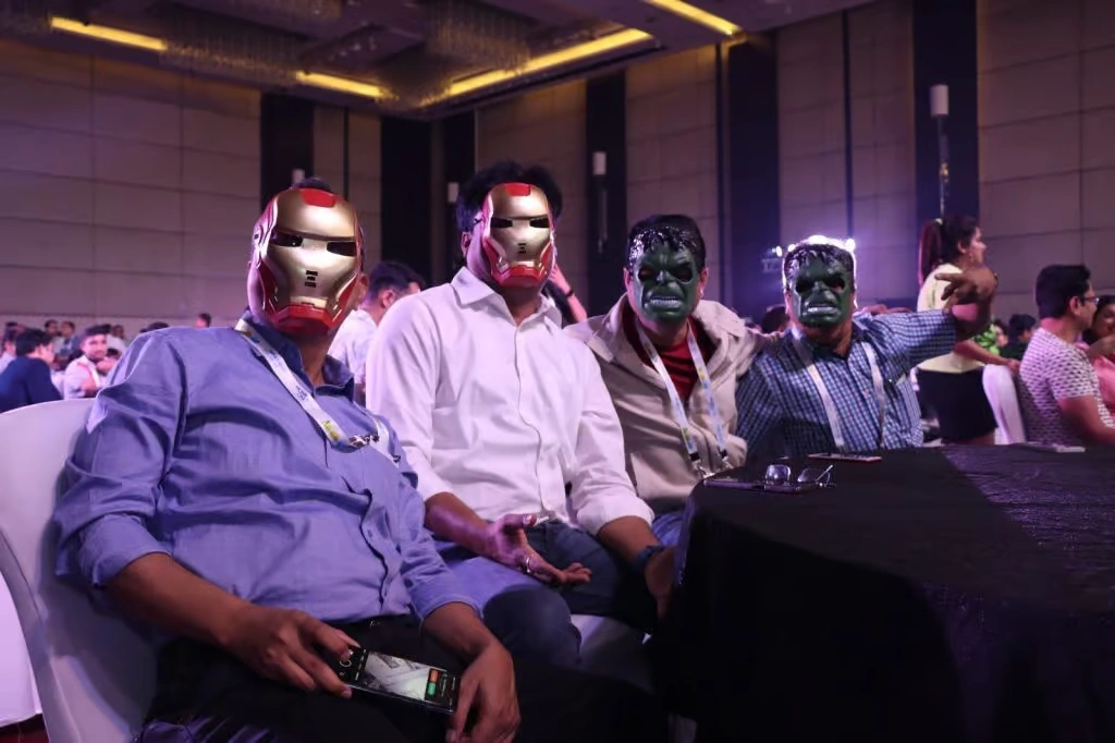 Four corporate employees sitting at a banquet table during an indoor gala, wearing Marvel-themed masks including Iron Man and The Incredible Hulk.