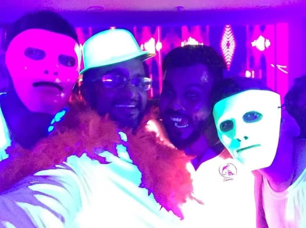 A high-energy corporate party scene under UV blacklights, featuring employees dancing and wearing glowing neon accessories, face paint, and LED glasses in a vibrant purple and blue environment.