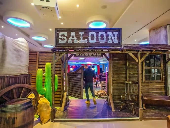A corporate event space decorated in a Wild West theme, featuring a large "Saloon" entrance with swinging wooden doors, cactus cutouts, wooden barrels, and warm, amber ambient lighting.