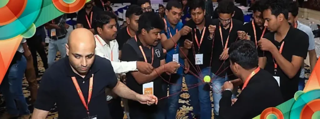 Building Stronger Teams: A Guide to Planning Essential Team Building Activities A group of corporate employees wearing black t-shirts and lanyards working together in a circle to balance a tennis ball using interconnected red strings.