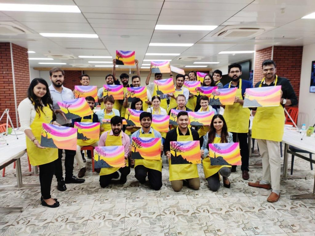 Paint-Social-Corporate-Team-Building-Creative-Kinship A large group of corporate employees in an office setting, wearing yellow aprons and holding up identical colorful landscape paintings they created during a team-building workshop.