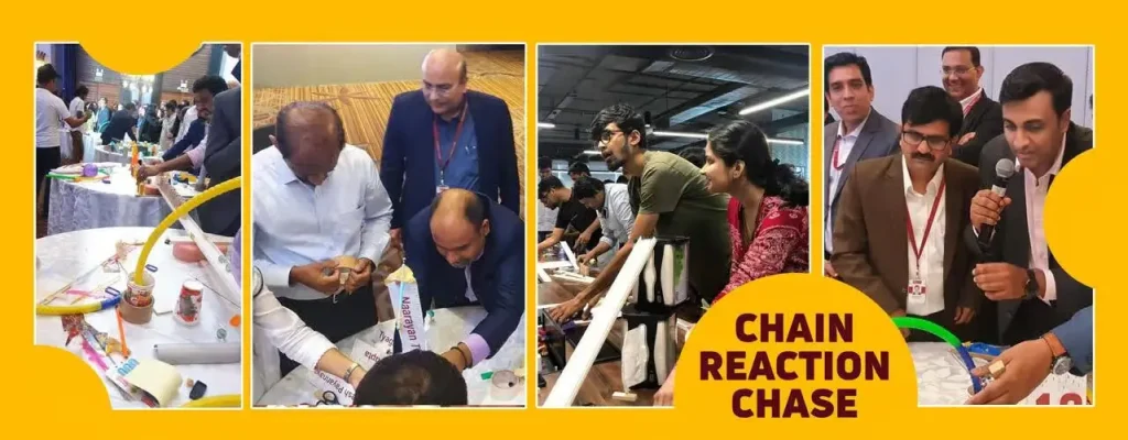 A wide-angle banner showing a diverse corporate team working together on the floor to build a complex Rube Goldberg-style chain reaction using pipes, balls, and wooden blocks.