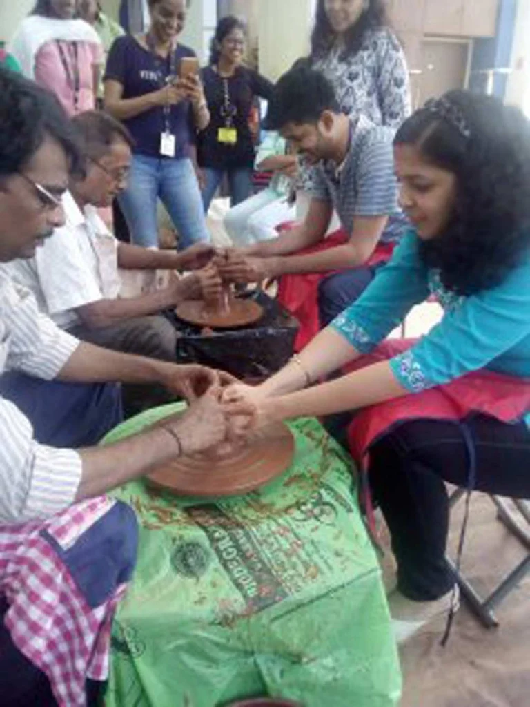 A group of corporate employees participating in a live pottery session, with a woman in a turquoise top focused on shaping clay on a potter's wheel under an instructor's guidance.
