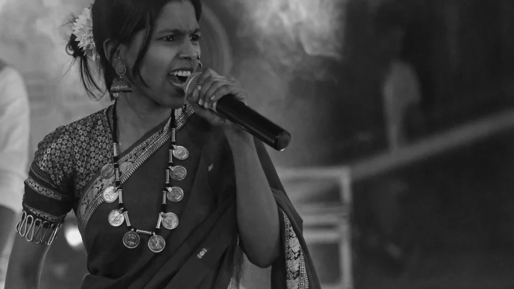 A black and white close-up of a female performer in traditional Indian attire and jewelry, passionately singing or speaking into a microphone.