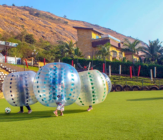 bubble-soccer-della-adventure-park-lonavala Corporate teams playing Bubble Soccer on a green turf at Della Adventure Park, Lonavala, with hills and luxury villas in the background.