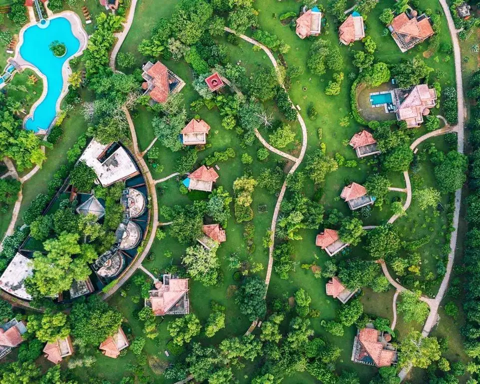 High-angle aerial view of Westin Sohna Resort & Spa, showing red-roofed villas nestled among lush green trees and winding pathways, featuring a bright blue swimming pool—a premium corporate retreat in the Aravali hills.