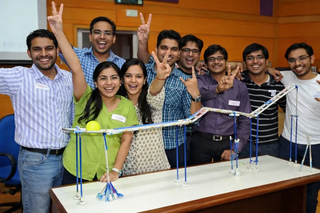 Roller Coaster Challenge - Team Alignment Success A diverse group of young corporate professionals cheering and posing with peace signs behind a large, handmade track model during a Roller Coaster Challenge team-building activity.