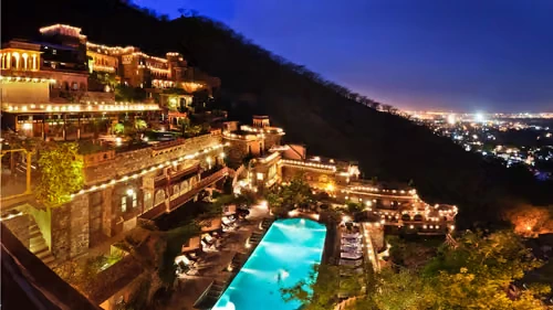 A breathtaking night view of the illuminated Neemrana Fort-Palace, showcasing its tiered stone ramparts, multiple courtyards, and a glowing turquoise swimming pool set against a dark mountain backdrop.