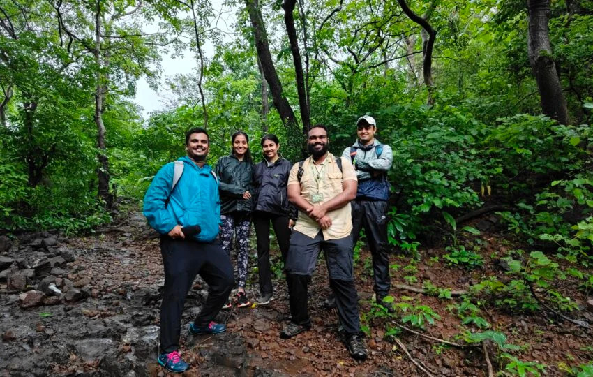 aarey-colony-forest-trail-mumbai-team-building A group of corporate professionals in hiking gear smiling on a lush, green forest trail in Aarey Colony, Mumbai, led by a professional guide.