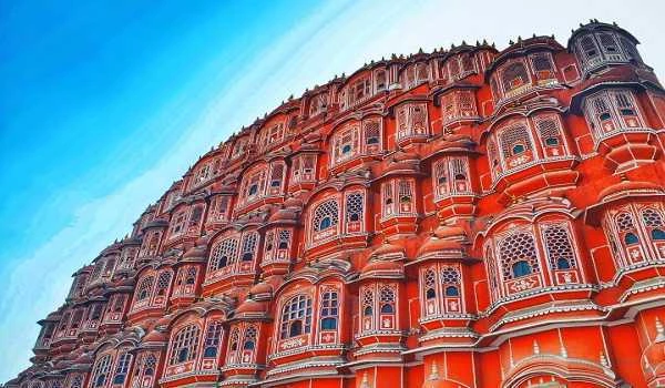 Close-up architectural view of the Hawa Mahal (Palace of Winds) in Jaipur, featuring its iconic pink sandstone facade, intricate honeycomb windows (jharokhas), and traditional Rajasthani craftsmanship against a clear blue sky.
