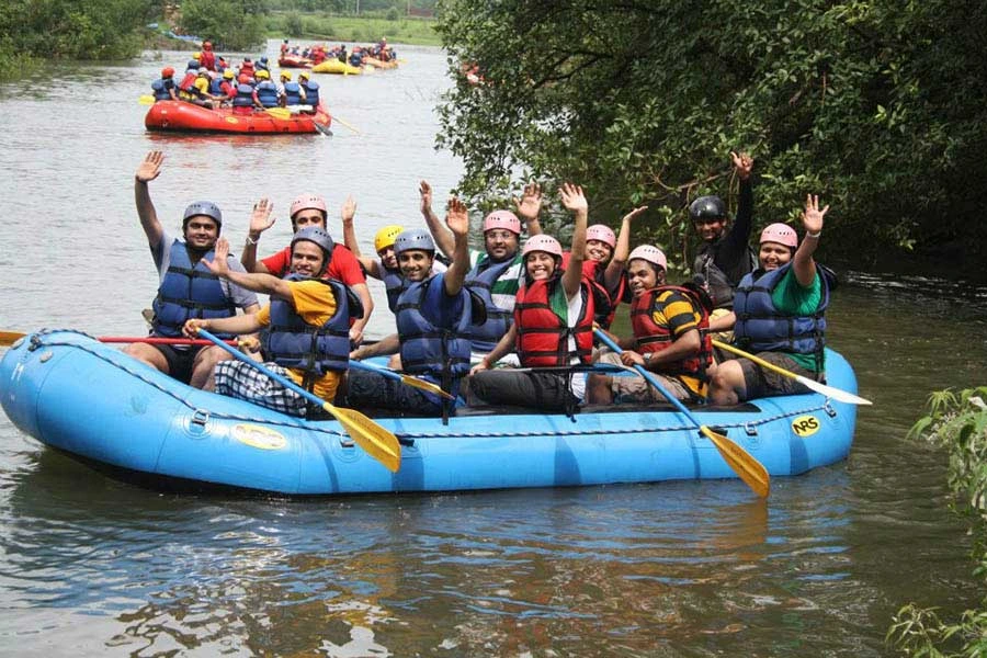 River-Rafting-in-Kolad-Kundalika-River A corporate team in blue life jackets and helmets cheering on a bright blue raft during a white-water rafting session on the Kundalika River in Kolad.