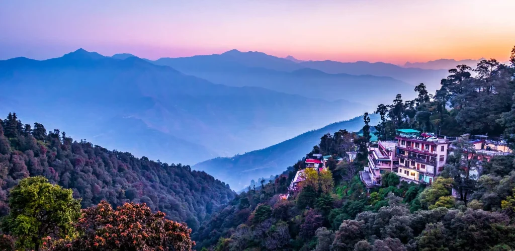 A wide panoramic view of Mussoorie at sunset, featuring multi-story buildings perched on steep green hillsides overlooking a vast valley of misty blue mountain ranges under a purple and orange sky.