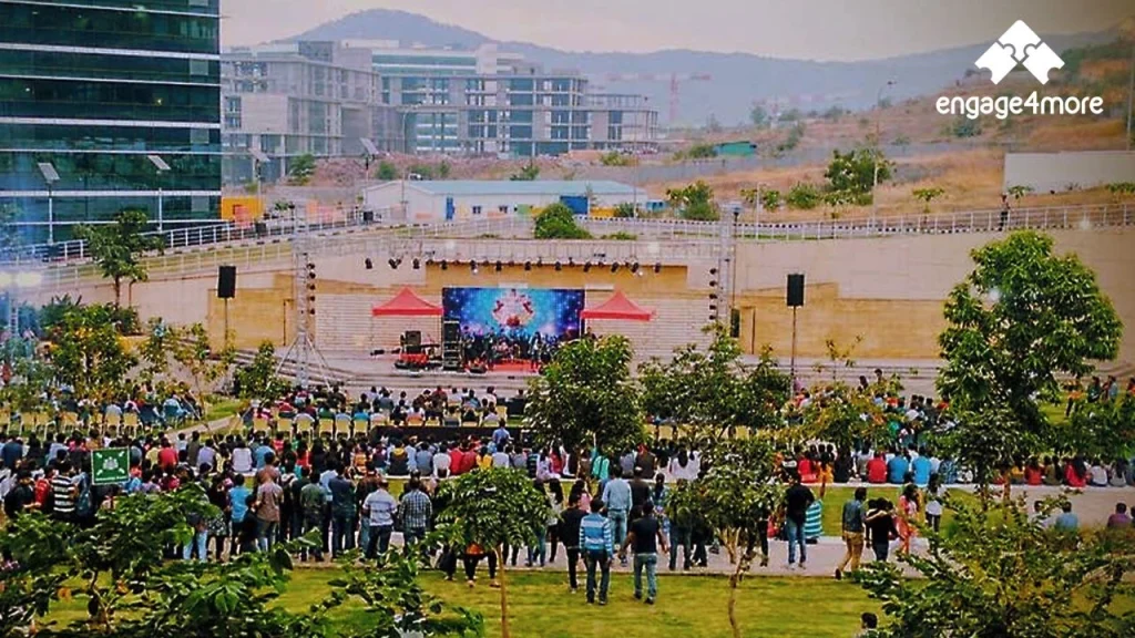 Why engage4more is the Top Team Building Company in Mumbai: 5 Hidden Adventure Hotspots for 2026 A wide-angle view of a large corporate outdoor event at an office park in Mumbai, featuring an engage4more stage, hundreds of attendees, and modern glass buildings in the background.