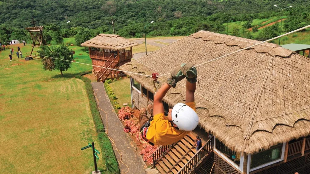 A corporate professional wearing a safety helmet and harness ziplining over a lush green meadow at a premium adventure resort near Pune