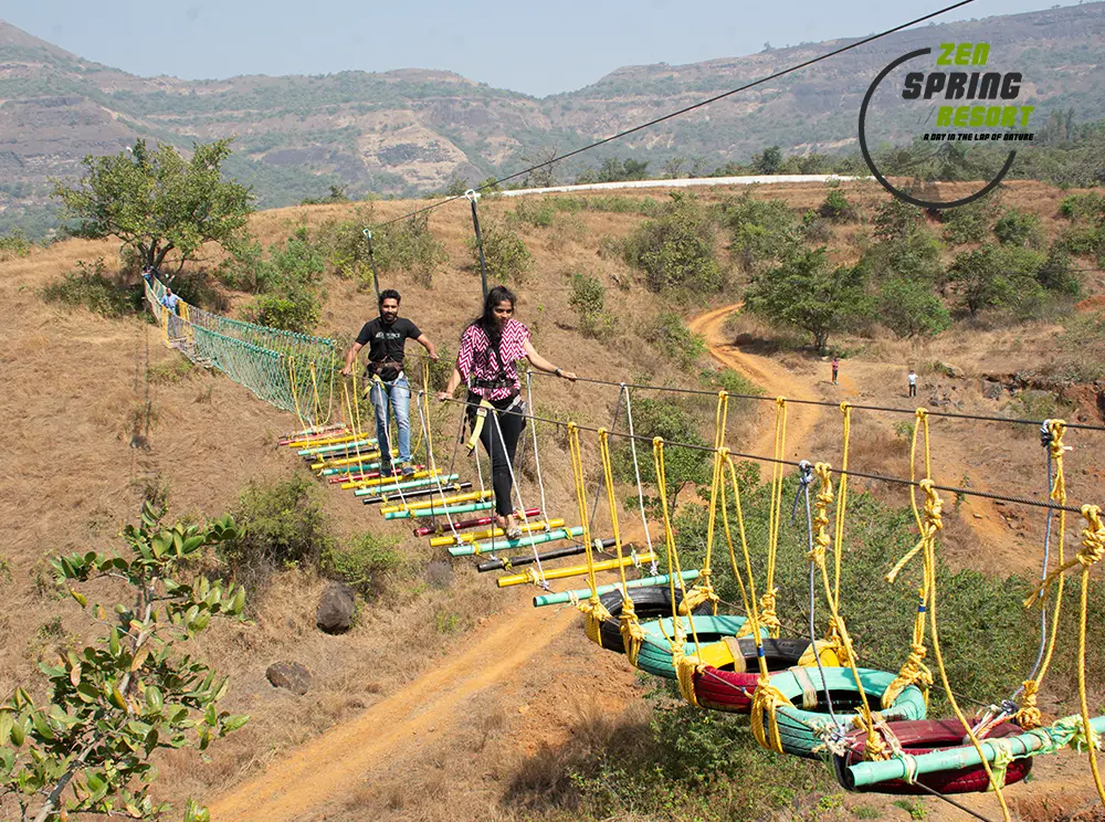 mulshi-leadership-retreat-zen-strategy-sanctuary Two corporate professionals carefully crossing a high-altitude suspension bridge made of ropes and colorful planks at Zen Spring Resort in Mulshi, with a vast mountain landscape in the background.