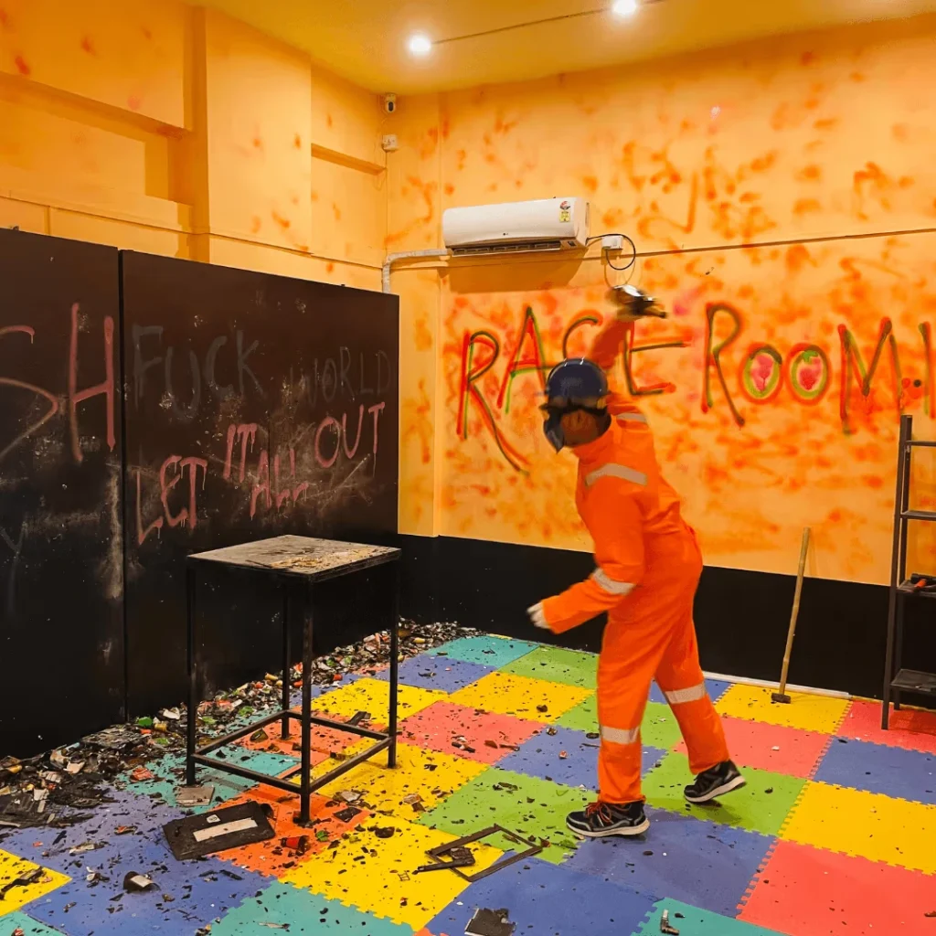 rage-room-mumbai-corporate-stress-relief-engagement A corporate professional in full safety gear and an orange jumpsuit using a sledgehammer to smash objects in a brightly colored Mumbai rage room with graffiti walls.