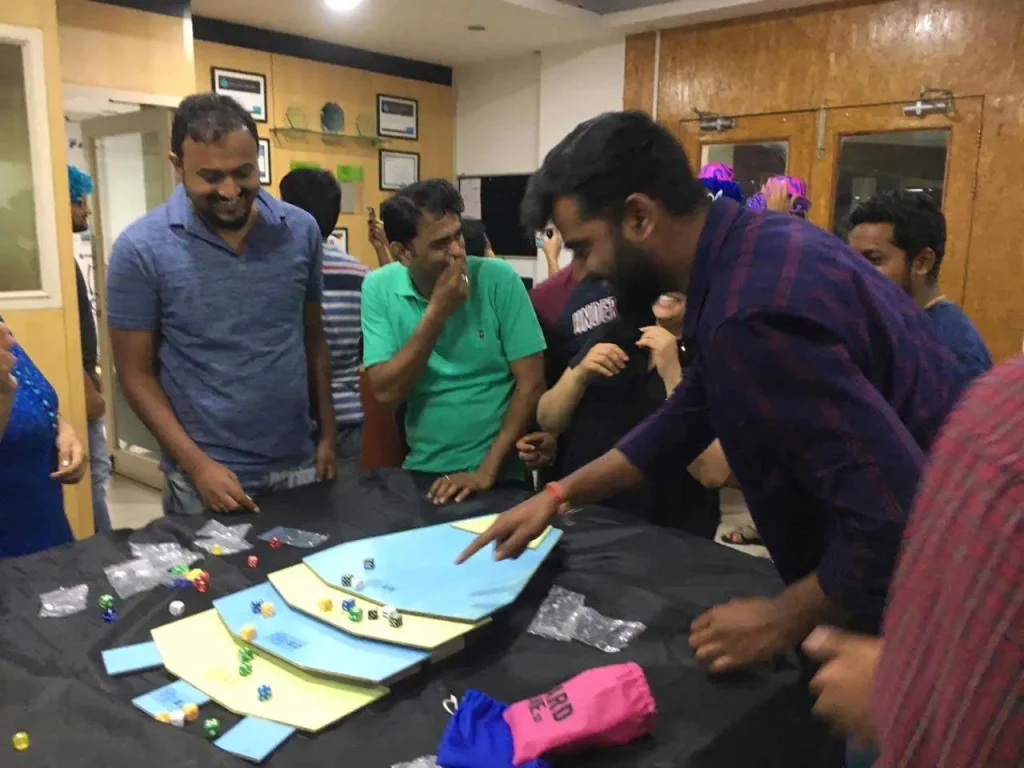 Corporate employees in a playful indoor setting engaged in a tabletop board game activity involving dice and colorful tokens.