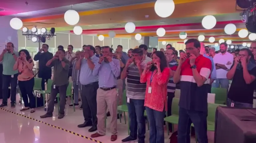 harmonica-jamming-corporate-team-building-mumbai A large group of corporate employees standing in an auditorium, all playing harmonicas together during a synchronized musical team-building session.