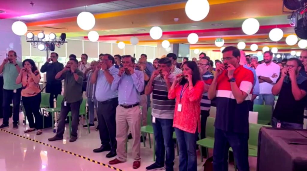 A large group of corporate employees participating in a synchronized harmonica jamming session in a brightly lit hotel ballroom in Kovalam.